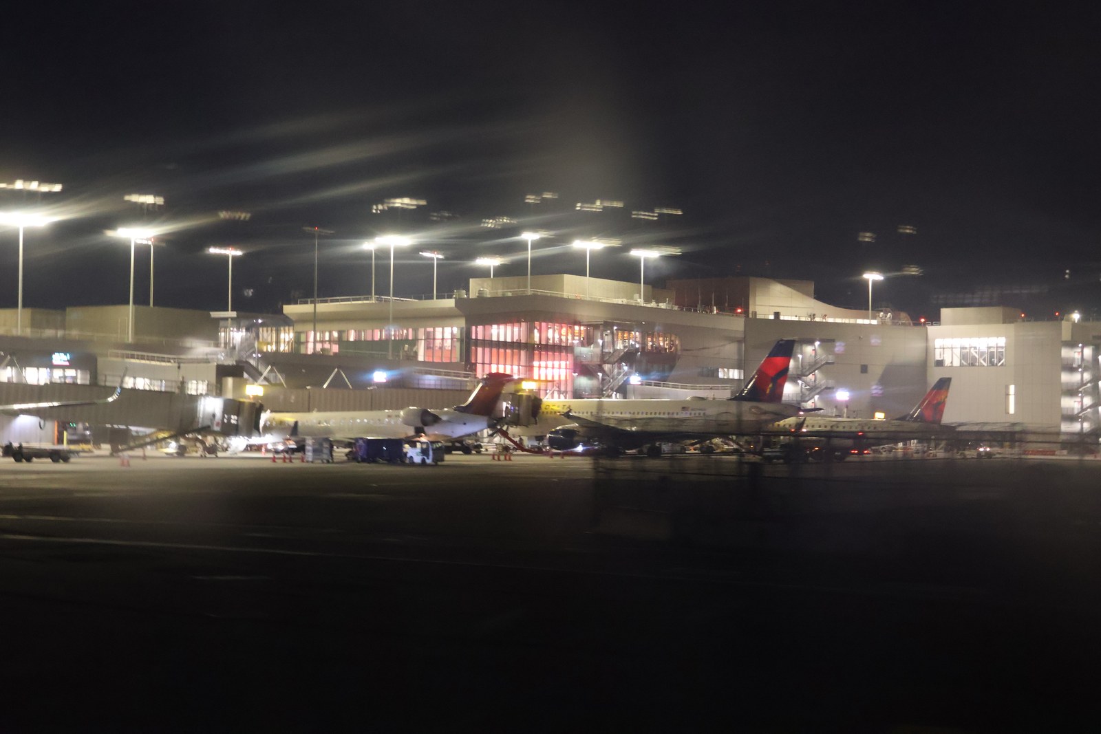 Night photograph taken from a taxiing aircraft at apron level of the Terminal C concourse in October 2023, with Delta aircraft parked at jet bridges, ground equipment, and the lit concourse windows behind